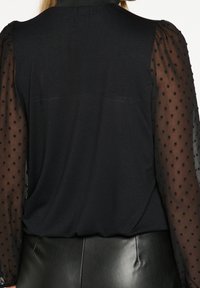 Top nero con un corpo in tessuto solido e maniche a sbuffo semitrasparenti decorate con pois neri testurizzati. Indossato con pantaloni in pelle nera.