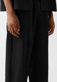 Närbild av en person klädd i lösa svarta byxor med händerna avslappnat vid sidorna mot en enkel ljus bakgrund.