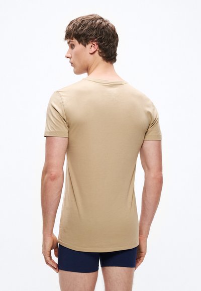 Μπεζ κοντομάνικη μπλούζα με στρογγυλή λαιμόκοψη και εφαρμοστή γραμμή, κατασκευασμένη από απαλό ύφασμα, εμφανίζεται από την πίσω πλευρά, συνδυασμένη με σκούρες μπλε βερμούδες.