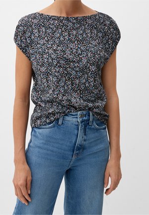 Frau trägt eine schwarze Bluse mit kurzen Ärmeln und pinken, weißen sowie blauen Sprenkeln, in hoch taillierte hellblaue Jeans gesteckt.
