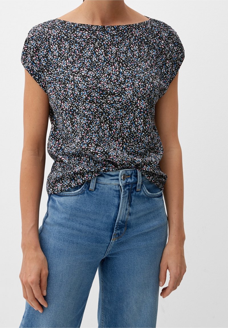 Femme portant un chemisier noir à manches courtes avec des taches roses, blanches et bleues, rentrée dans un jean bleu clair taille haute.