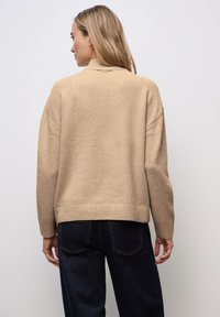 Beżowy sweter dzianinowy z wysokim dekoltem, luźnym krojem, prążkowanym dołem i opuszczonymi ramionami. W połączeniu z ciemnoniebieskimi jeansami o gładkiej teksturze.