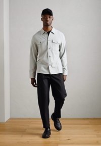 Ανοιχτό γκρι πουκάμισο με κουμπιά, δύο τσέπες στο στήθος και μακριά μανίκια, συνδυασμένο με μαύρες φαρδιές παντελόνες και μαύρες παπούτσια. Το μοντέλο φοράει ένα μαύρο καπέλο.