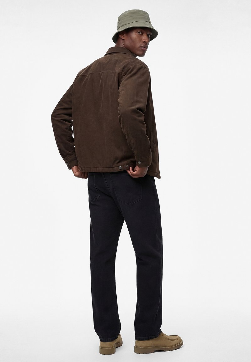 Man wearing olive bucket hat, brown jacket, black pants, and tan boots, standing and looking over shoulder against plain background.