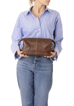 Donna con camicia a righe blu e jeans che tiene una borsa per cosmetici in pelle marrone con cerniera e manici su ciascun lato.