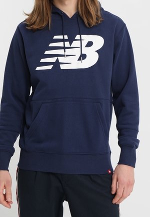 Personne portant un sweat à capuche bleu marine avec un grand logo blanc "NB" et un pantalon noir avec des bandes latérales, les mains détendues sur les côtés.