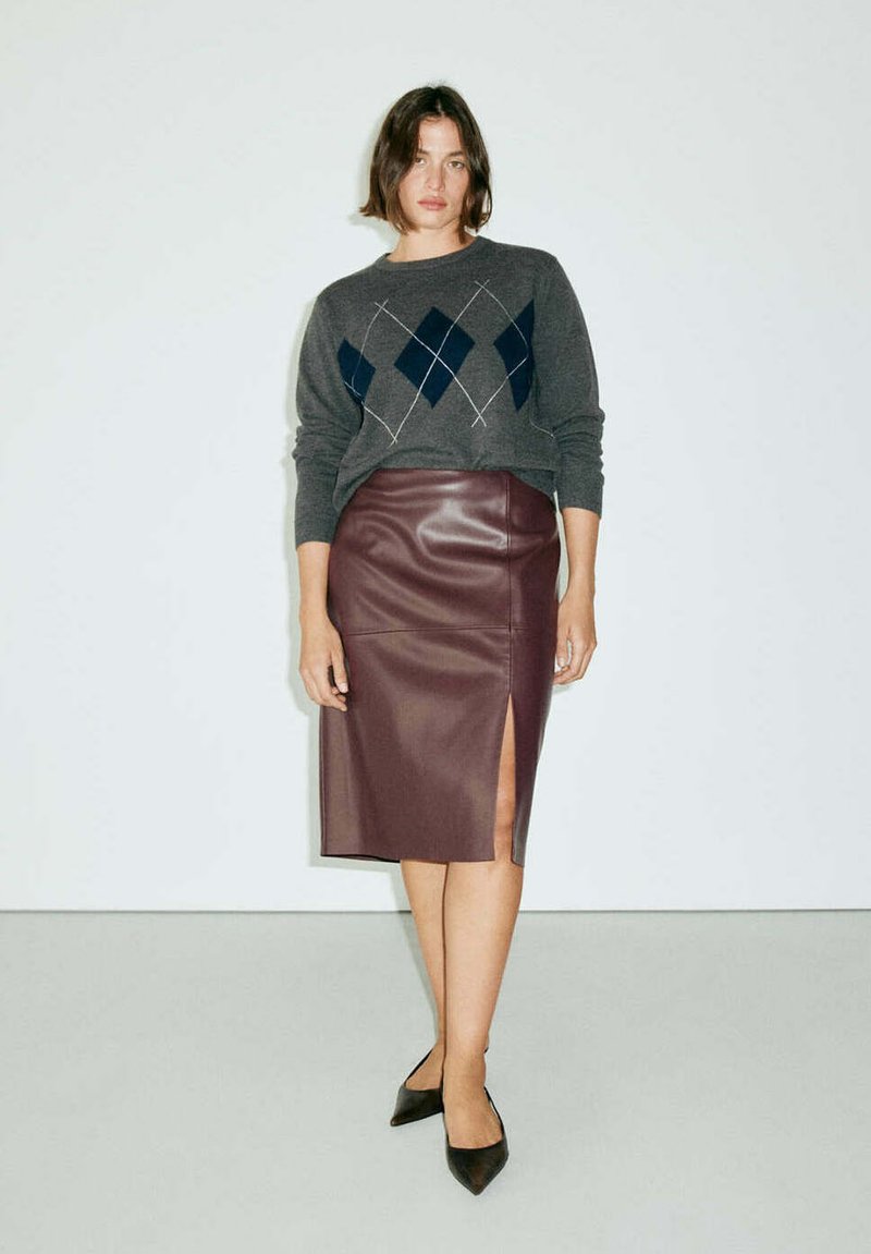 Femme portant un pull gris à motif losanges et une jupe en cuir marron longueur genou avec une fente devant, debout devant un fond blanc uni.