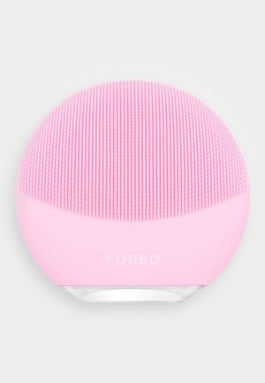 Różowe silikonowe urządzenie do oczyszczania twarzy o zaokrąglonym kształcie, z teksturowaną powierzchnią składającą się z krótkich i długich włosków, oznaczone napisem "FOREO."