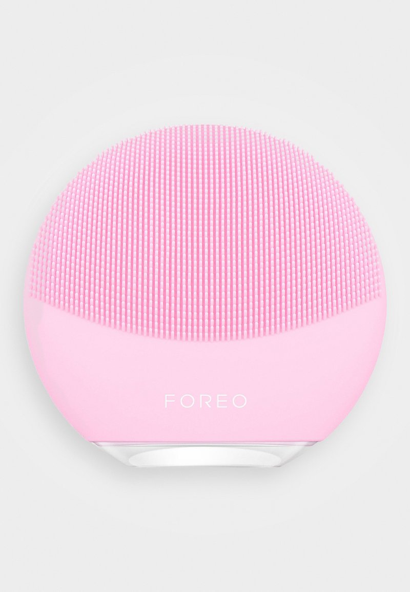 Dispositif de nettoyage facial en silicone rose avec une forme arrondie, dotée d'une surface texturée de poils courts et longs, étiqueté "FOREO."