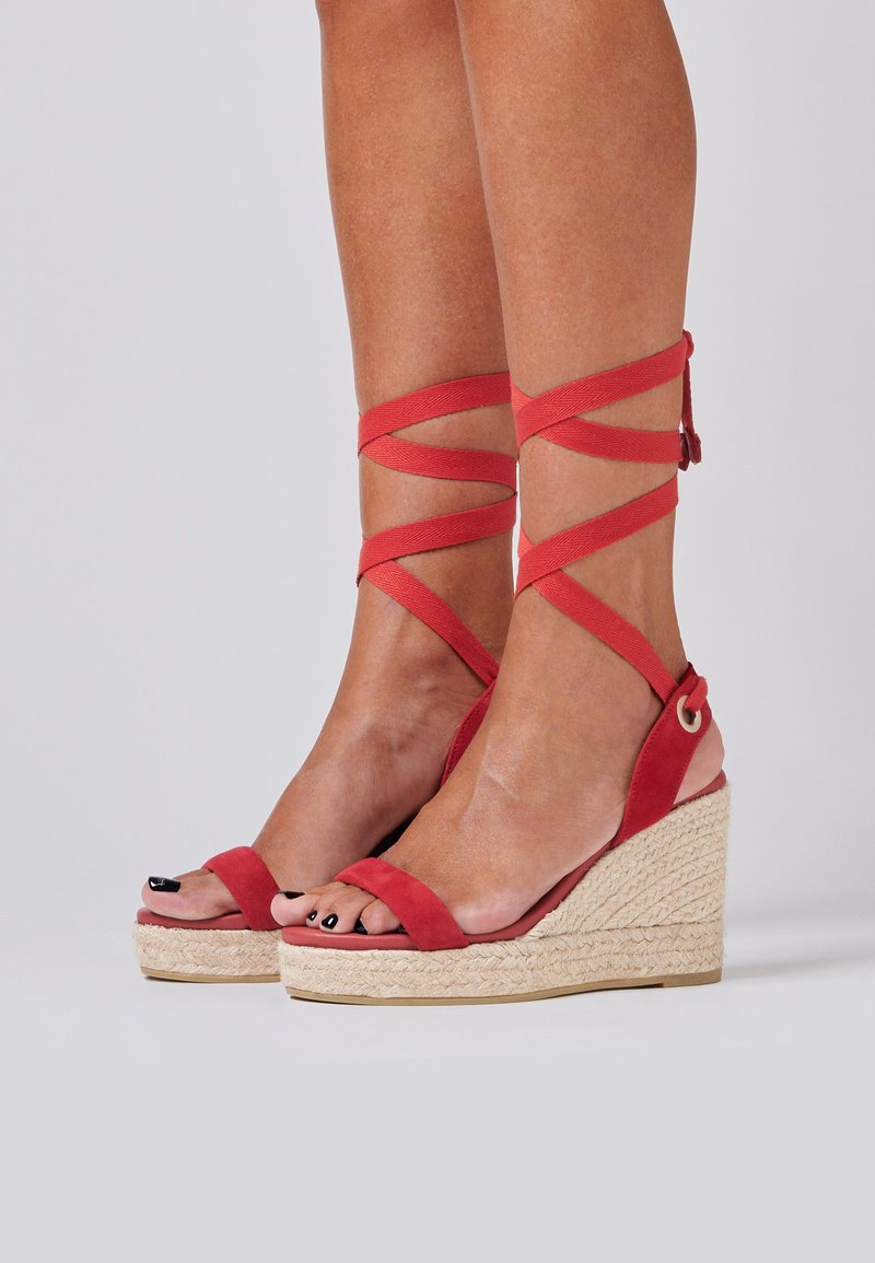 Red suede wedge sandals with crisscross ankle straps, jute-wrapped platform, and open toes. Features a rounded shape and minimal hardware.