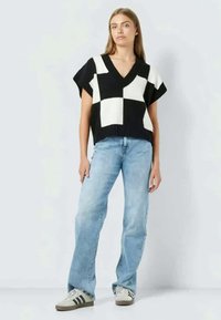 Melnā un baltā šaha raksta adīta tops ar V-veida kakla izgriezumu, apvienots ar gaiši ziliem taisnās kājas džinsiem un pelēkiem sporta apaviem.