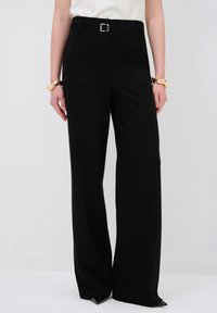 Schwarze weite Hosen aus glattem Stoff, mit einem schmalen Bund und einem Gürtel. Der Saum ist leicht verjüngt und endet über spitzen Schuhen.