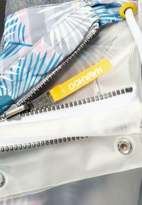 Mångficka väska med en transparent plastinteriör, grått tygutsidan med ett bladmönster och en gul etikett. Silverdragkedjedetalj.