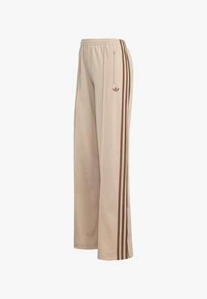 Pantalones deportivos beige con triple raya lateral marrón, cintura elástica, bolsillo con cremallera y logo de trébol de Adidas en el muslo derecho.