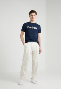Navy modrá bavlněná tričko s bílým logem "Barbour." v kombinaci s krémovými skládacími kalhotami a bílými teniskami, na neutrálním světlém pozadí.