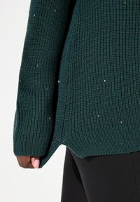 Mörkgrön stickad tröja med ribbad textur, med en sidoklyfta och subtila metalliska accenter. Långa ärmar visas.