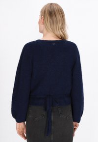 Blonde hajú nő, aki sötétkék, bolyhos pulóvert visel, derékban megkötve, és sötétszürke farmer szoknyát, hátulról látható.