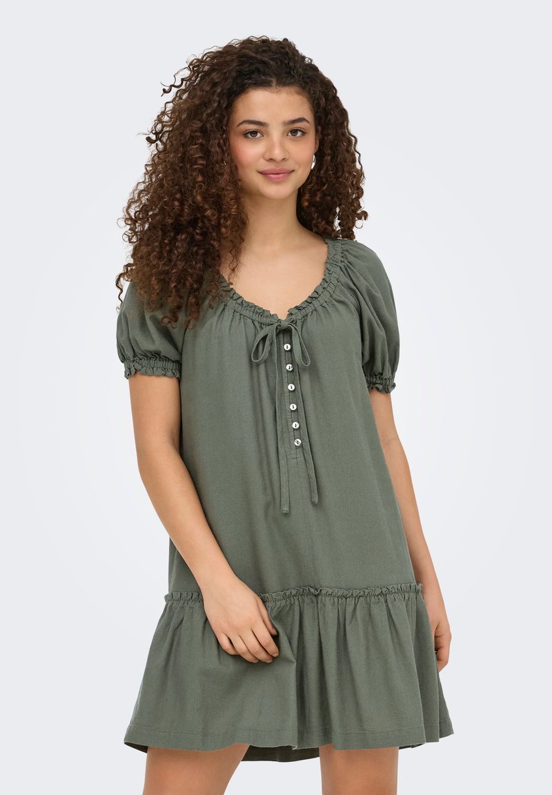 Jeune femme aux cheveux bouclés portant une robe olive à manches courtes boutonnée avec un ourlet volanté sur fond neutre.