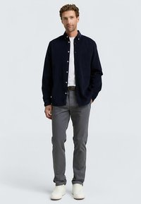 Marineblauw overhemd met knopen, grijze broek en witte sneakers. Het overhemd heeft een zak en is gemaakt van een textuurstof, wat een gelaagde uitstraling creëert.