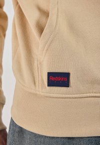 Sweat-shirt beige avec une finition côtelée et une poche avant, arborant une étiquette bleu marine avec le texte rouge "Redskins 1984" près de la taille.