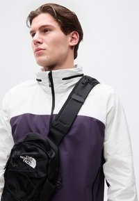 Νεαρός άνδρας που φοράει λευκό και μωβ μπουφάν με φερμουάρ και μαύρη τσάντα χιαστί σε λευκό φόντο.