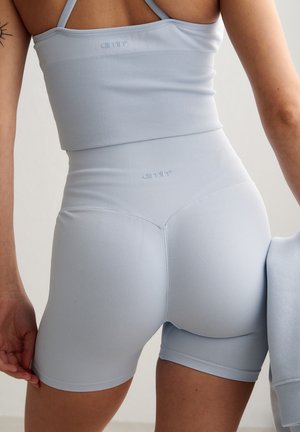 Kvinne iført lys blå sømløs ribbestrikket sports-BH og høytlivs biker-shorts, holder matchende hettejakke, mot ensfarget bakgrunn.