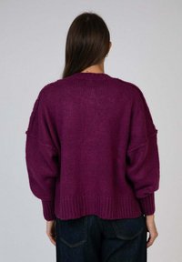 Femme avec de longs cheveux bruns portant un pull violet foncé à côtes lâches et un jean bleu foncé, vue de dos contre un fond uni.