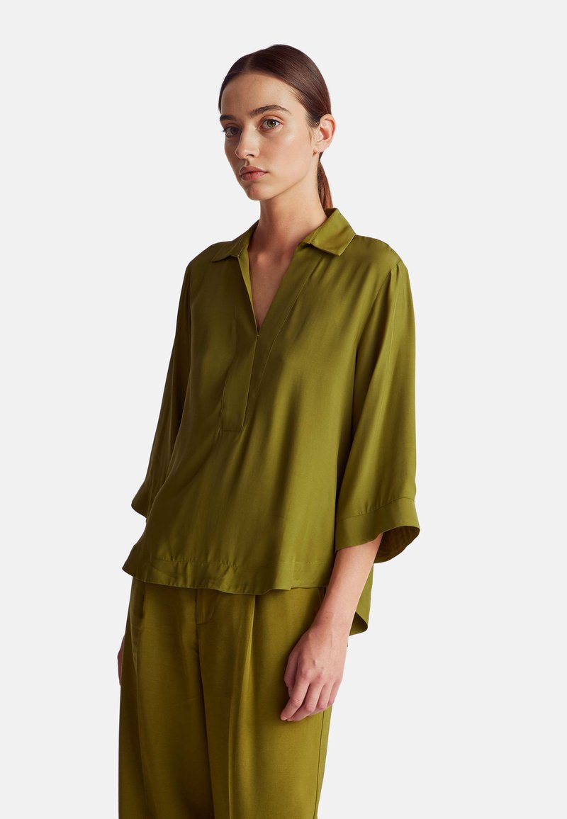 Blusa leggera di colore verde oliva, con scollatura a V, maniche a tre quarti e una vestibilità ampia. Tessuto liscio con una leggera lucentezza.