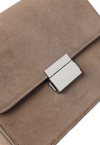 Vaaleabeige mokkanahkalaukku, jossa on sileä ja pehmeä pinta, hopeinen metallilukko ja kohokuvioitu logo. Kompakti ja suorakaiteen muotoinen muotoilu.