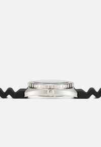 Montre en acier inoxydable argentée avec un bracelet en caoutchouc noir texturé, présentant une lunette nervurée et un verre rond lisse.