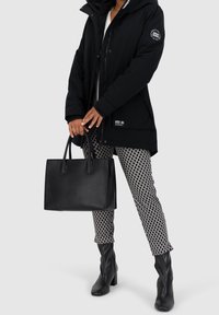 Černá parka s kapucí, zipem a patentním uzávěrem, černé kalhoty s potiskem a černá kožená kabelka. Černé kozačky na vysokém podpatku ke kolenům.