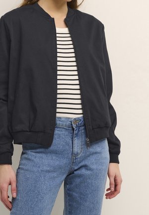 Personne portant une veste noire zippée par-dessus une chemise rayée blanche et noire avec un jean bleu, debout devant un fond clair uni.