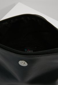 Czarna torba typu pouch z zamknięciem na zamek, cechująca się gładką powierzchnią, srebrnym zapięciem oraz miękkim czarnym wykończeniem wnętrza. Logo widoczne wewnątrz.