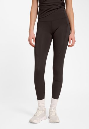 Vrouw draagt zwarte nauwsluitende leggings met subtiele gebogen naden, witte sokken en lichtbeige sneakers, staand tegen een effen achtergrond.