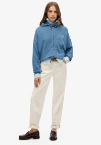 Felpa blu con zip, cappuccio, tasche frontali e coulisse, abbinata a pantaloni crema e mocassini scuri, che mostrano un look casual.