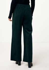 Breda byxor i kordfladdrig material i mörkgrön färg, med ett texturerat ribbat mönster, elastisk midja och en enda bakficka. Slät yta.