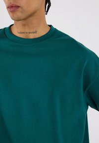 Türkiz pulóver, puha anyagból készült, klasszikus kerek nyakkal és sima textúrával, látható varrási részletekkel.