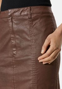 Brauner, taillierter Rock aus einem glatten, strukturierten Material mit seitlichen Taschen, sichtbaren Nähten und einem hohen Bund. Hand auf dem Stoff platziert.