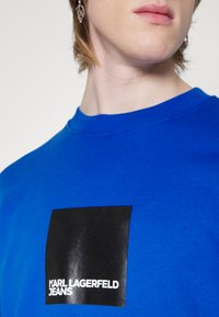 Plava pamučna majica s crnim kvadratnim logotipom i bijelim natpisom "KARL LAGERFELD JEANS" na lijevoj strani prsnog područja. S kratkim rukavima i okruglim izrezom.