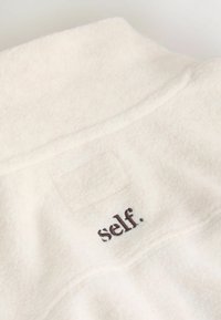 Weißes Fleece-Kleidungsstück mit hohem Kragen, das auf dem Rücken ein kleines, dunkelbraunes "self."-Logo trägt. Weiche Textur und schlichtes Design.