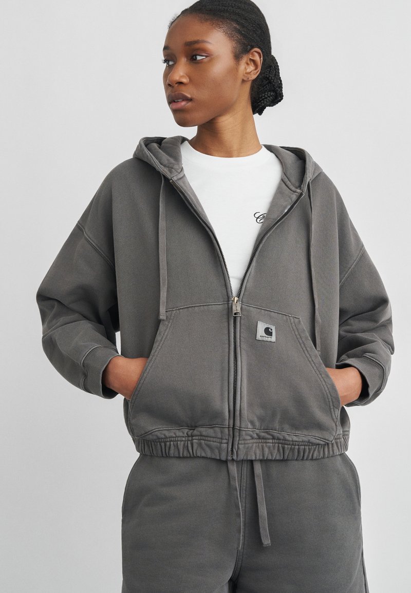 Donna con indosso una felpa grigia con zip e pantaloni della tuta coordinati con le mani nelle tasche, che guarda a sinistra su uno sfondo semplice.