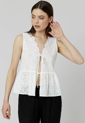 Blouse blanche sans manches avec une silhouette peplum, un col scallopé et un motif de fleurs en dentelle. Fermeture nouée à l'avant.