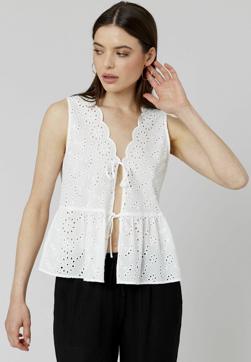 Blouse blanche sans manches avec une silhouette peplum, un col scallopé et un motif de fleurs en dentelle. Fermeture nouée à l'avant.