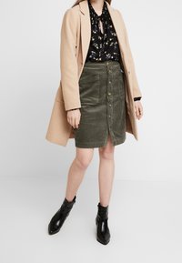 Un manteau beige clair porté sur un blouse noire à fleurs et une jupe en velours côtelé vert olive avec des boutons sur le devant et des poches. Bottines noires.