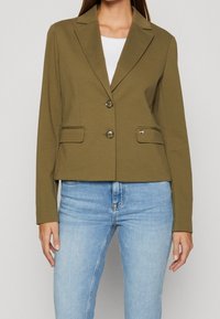 Žena v olivově zeleném saku se dvěma knoflíky a předními kapsami, oblečená přes bílé tričko, v kombinaci se světle modrými džíny.