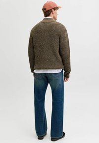 Brauner gestrickter Pullover mit gerippter Textur, kombiniert mit weit geschnittenen blauen Jeans. Das Modell trägt ein helles Hemd darunter.