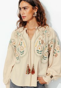 Blouse beige brodé avec des motifs floraux en vert, orange et jaune. Comprend des franges et des poignets froncés pour ajouter de la texture.