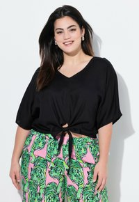 Top nero a V con maniche larghe, annodato sul davanti. Abbinato a pantaloni rosa con un motivo di tigrato verde. Tessuto liscio.