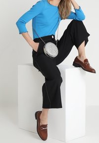 Blå långärmad topp, svarta vida byxor med vit sömnad, silverfärgad cirkulär väska med dekorationer, bruna loafers med spänne.