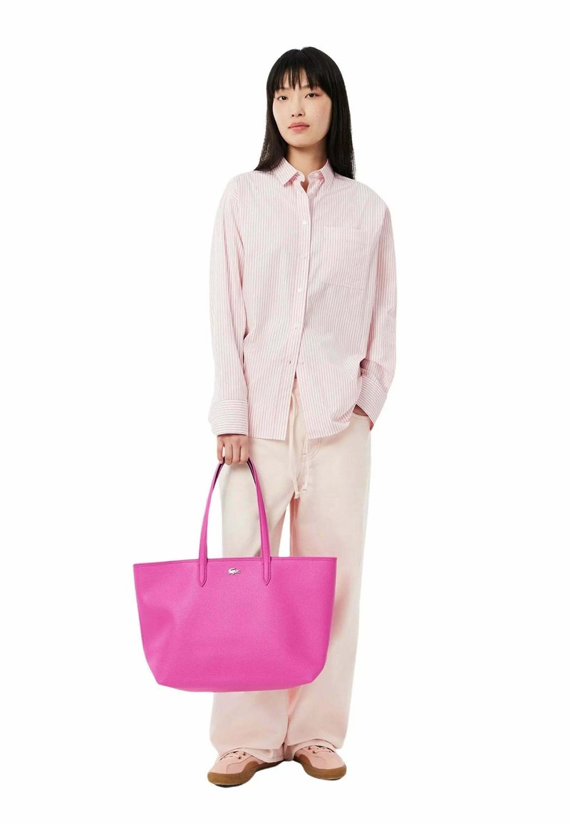 Sac fourre-tout rose texturé avec deux poignées, associé à une chemise rayée rose clair et un pantalon crème. Détail de logo simple sur le sac.